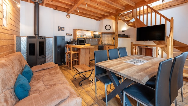 Interior acogedor de una cabaña con una mesa de comedor de madera, sillas, un sofá, una zona de cocina y un televisor montado en una viga de madera.