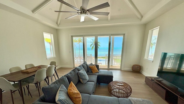 A spacious living room with a grey sectional sofa, a dining table, and a TV. Large windows offer a view of the beach and ocean. - Slipstream 2.0 - A Modern Beachfront Escape (Long Island)