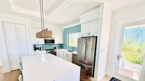A modern kitchen with white cabinetry, a stainless steel refrigerator, a large island with bar stools, and a view of greenery through the sliding glass door. - Slipstream 2.0 - A Modern Beachfront Escape (Long Island)