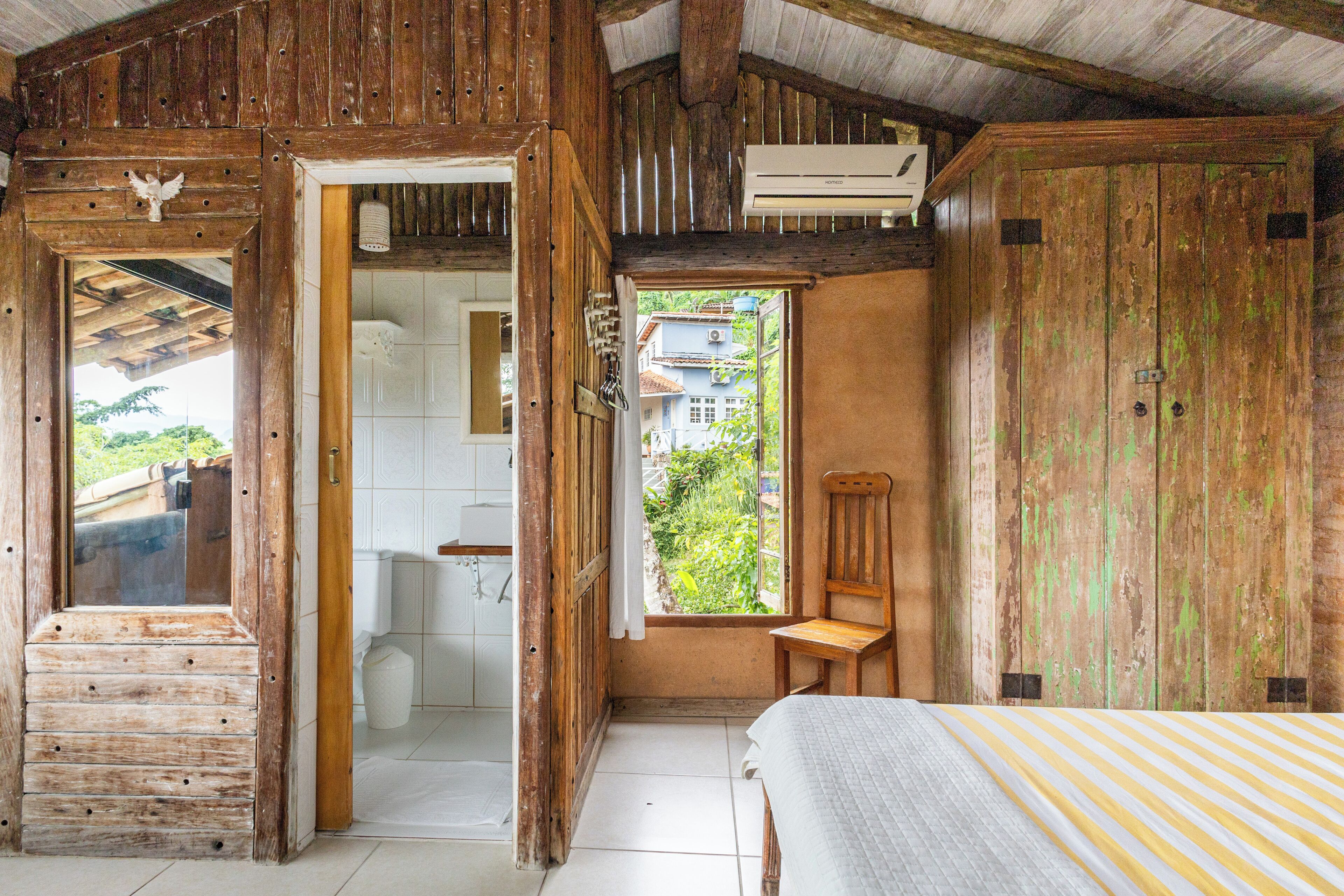Una habitación rústica de madera con una cama, un banco de madera y vista a una casa a través de una ventana.