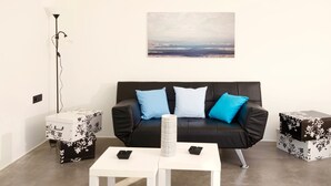 Uma sala de estar moderna com um sofá de couro preto, duas mesas laterais brancas e um grande quadro paisagístico na parede.