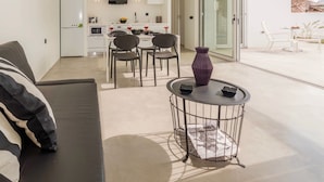 Um espaço de estar moderno com sofá, sala de jantar com cadeiras, cozinha com armários e eletrodomésticos, e uma grande porta de vidro que dá para área externa.