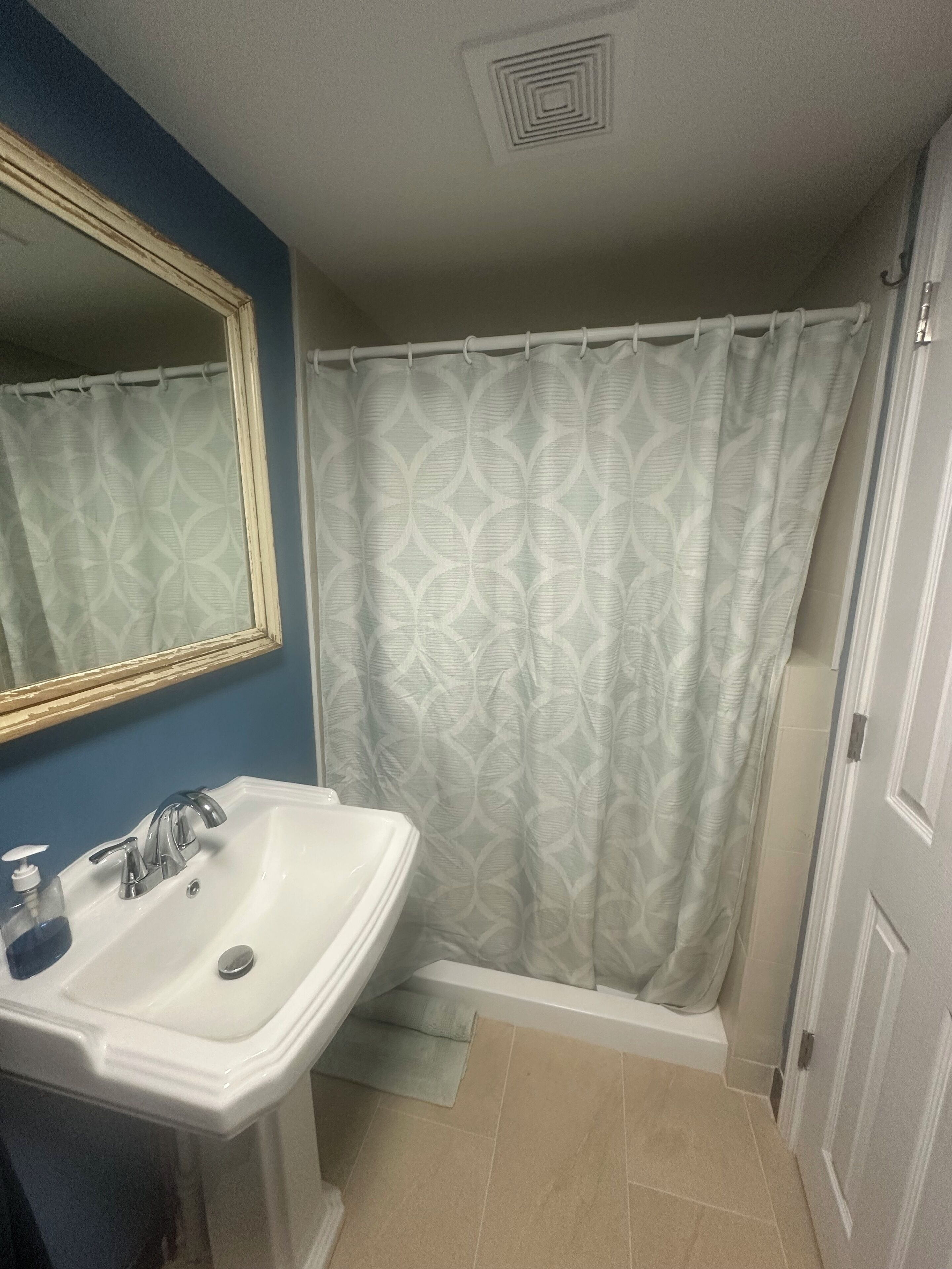 Un baño con lavamanos blanco, espejo, cortina de ducha estampada con hojas y azulejos color beige.