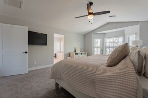 Une chambre avec un grand lit, un ventilateur de plafond, un téléviseur fixé au mur et une fenêtre avec des stores.
