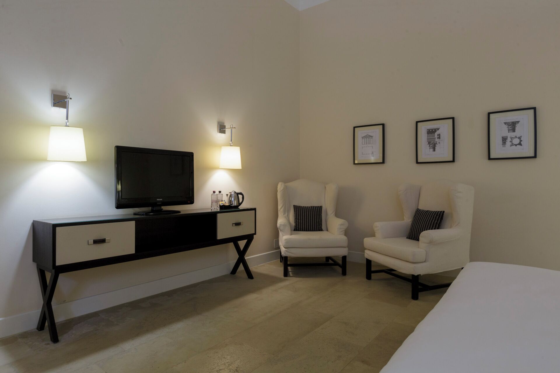 A hotel room with a bed, a desk with a TV, two armchairs, and wall-mounted lamps.