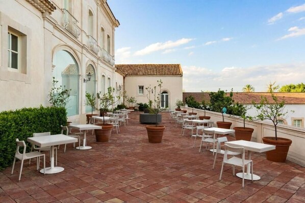 An outdoor patio with white tables and chairs, surrounded by potted plants and a building with arched windows. - Hotiday Siracusa Floridia (Syracuse)