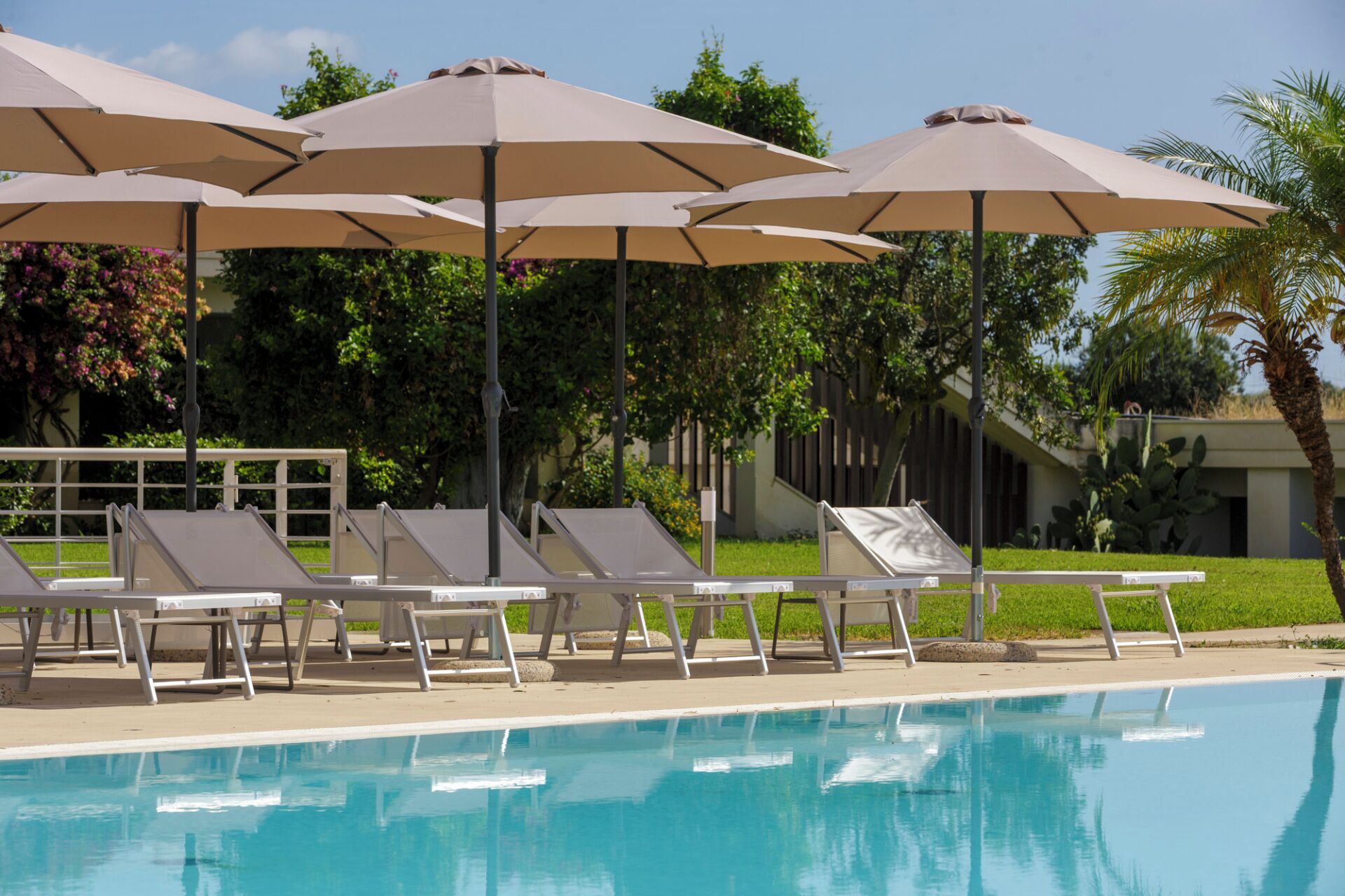 Zona de piscina con sillones de descanso y sombrillas, rodeada de vegetación y un edificio al fondo.