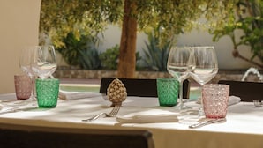 Mesa lista para comer, con copas de vino y cristalería decorada, frente a un fondo de follaje y un árbol.