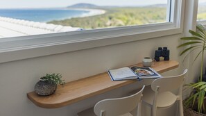 Ein Holztisch mit einem Buch, einer Tasse und Ferngläsern vor einem Fenster mit Blick auf eine Küstenlandschaft.