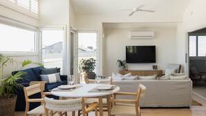 Een ruime woonkamer met een bank, eettafel en stoelen, een flatscreen tv aan de muur bevestigd, en grote ramen met uitzicht op buiten.