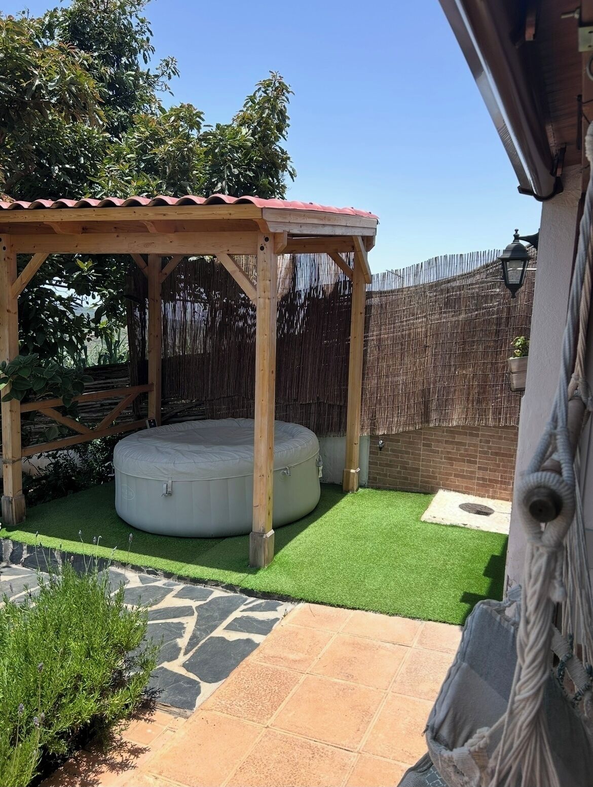 Un pabellón de madera con techo rojo, un colchón inflable redondo, césped artificial y un patio con baldosas de piedra.