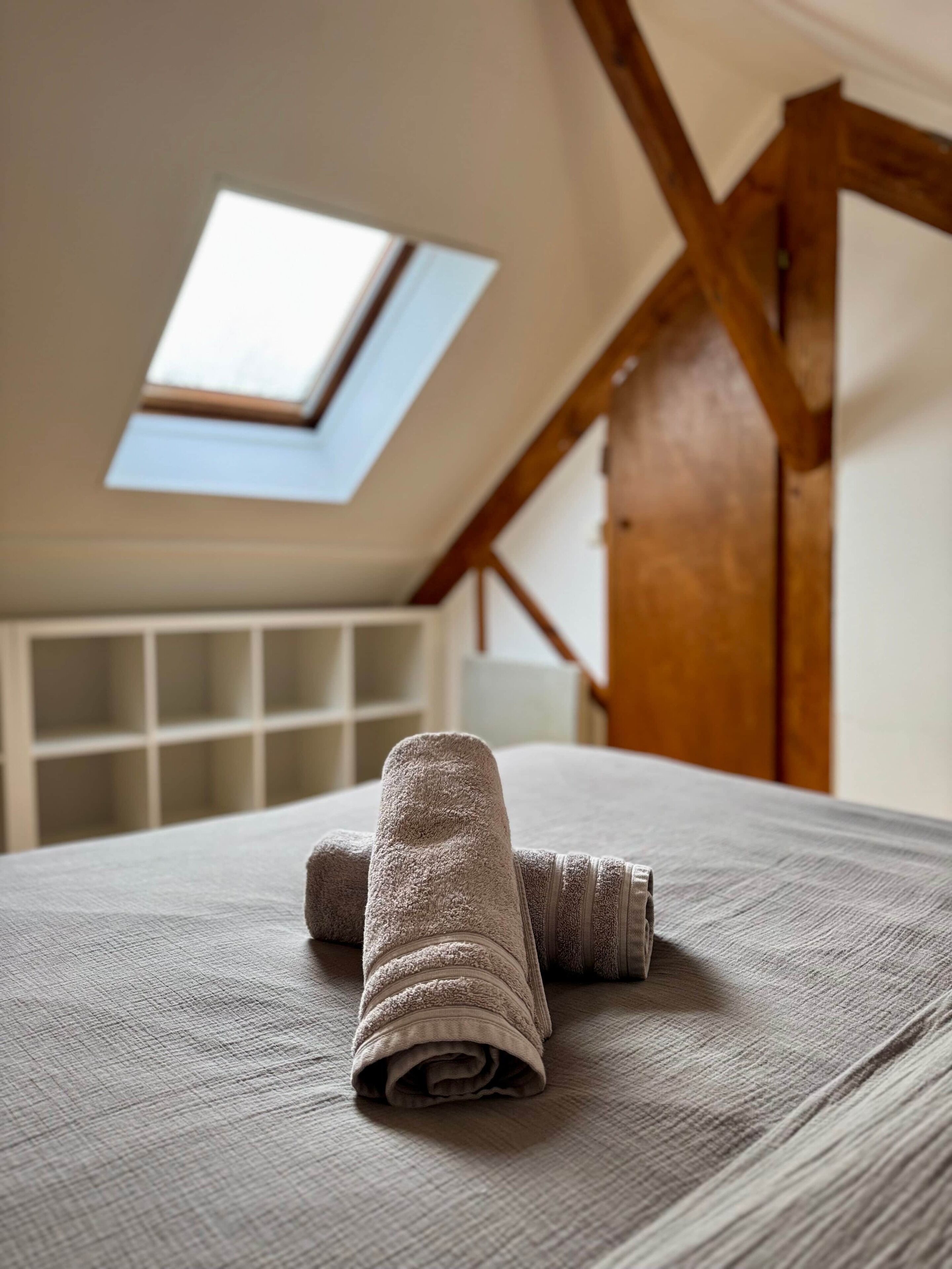 Ein Zimmer mit Dachfenster, einem Bett mit zusammengelegtem Handtuch und einer Holztür.