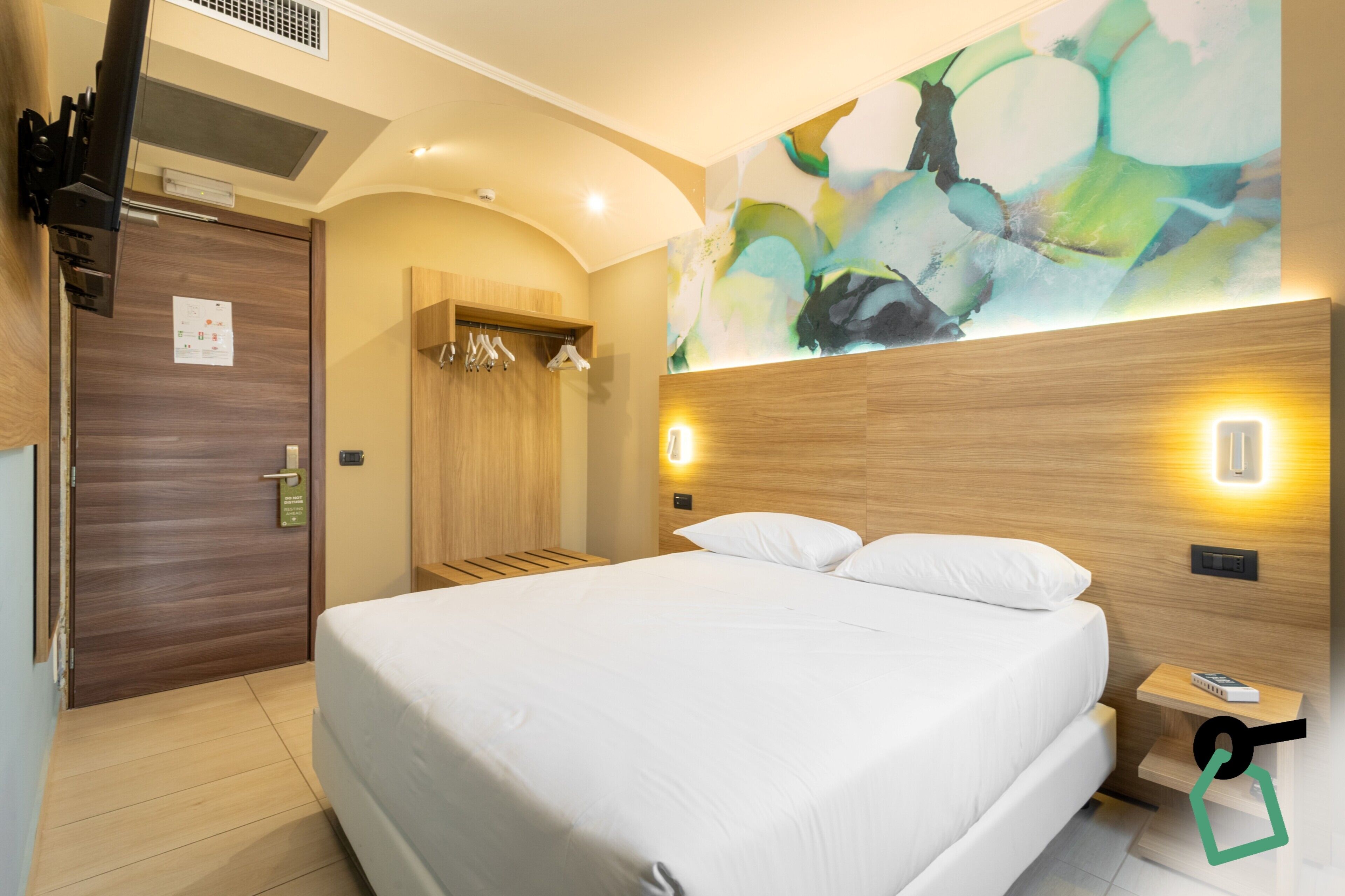 A modern hotel room with a large bed, wooden headboard, abstract wall art, and a built-in wardrobe.