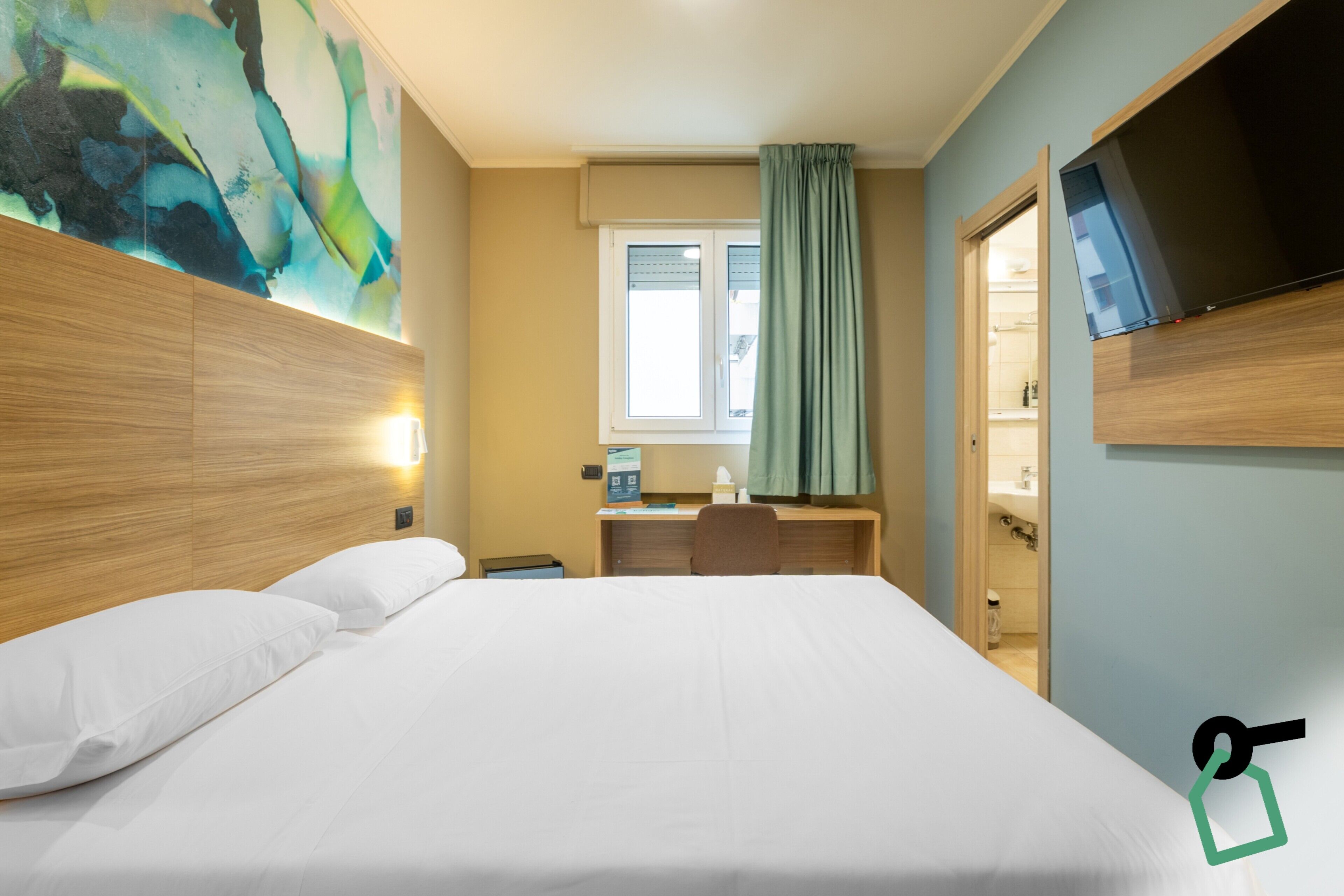 Habitación de hotel con una cama grande, un escritorio con silla, televisor montado en la pared y una ventana con cortinas.