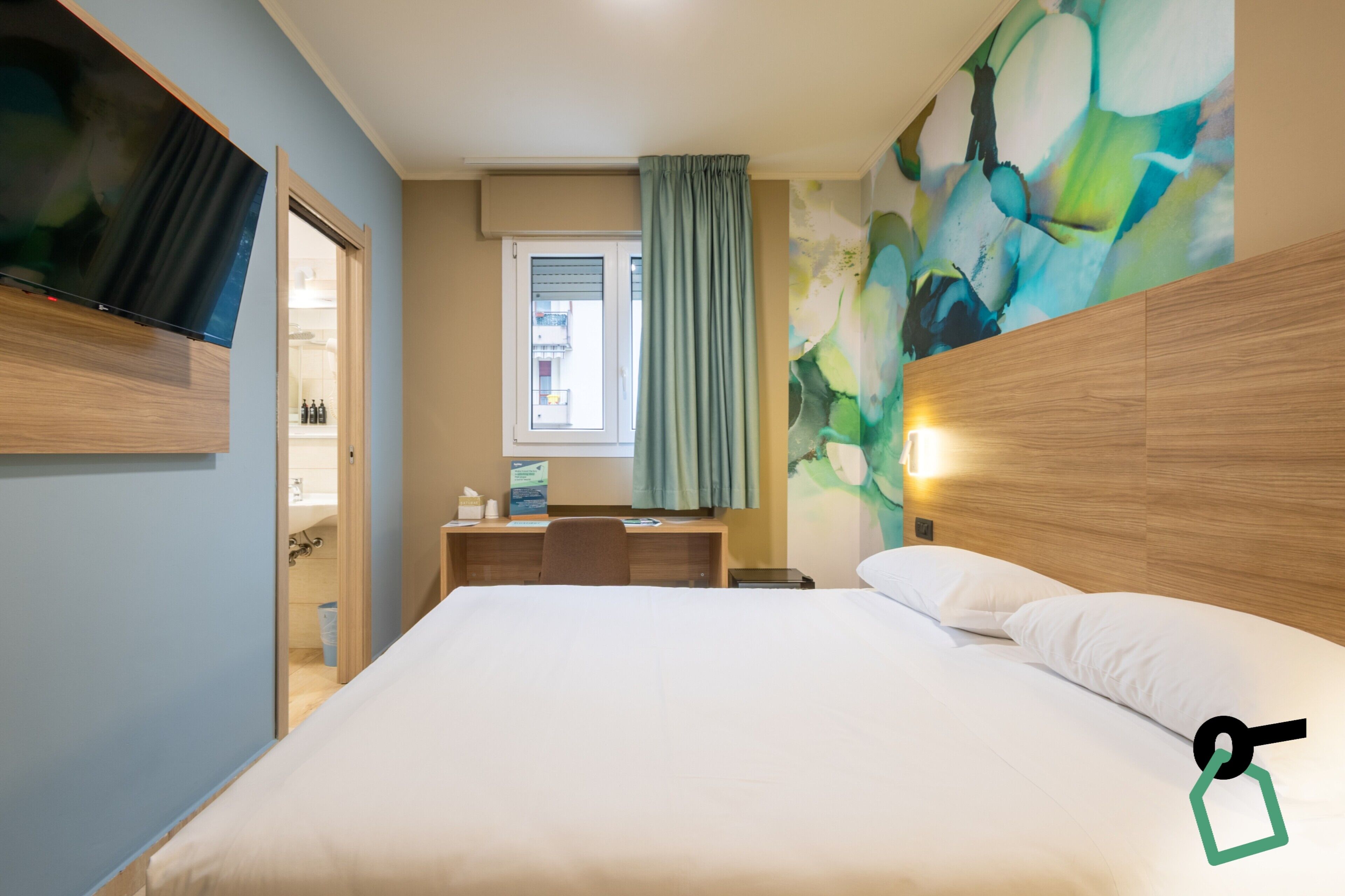 A hotel room with a bed, a desk, a chair, a TV mounted on the wall, a window with curtains, and a wall with a colorful abstract painting.