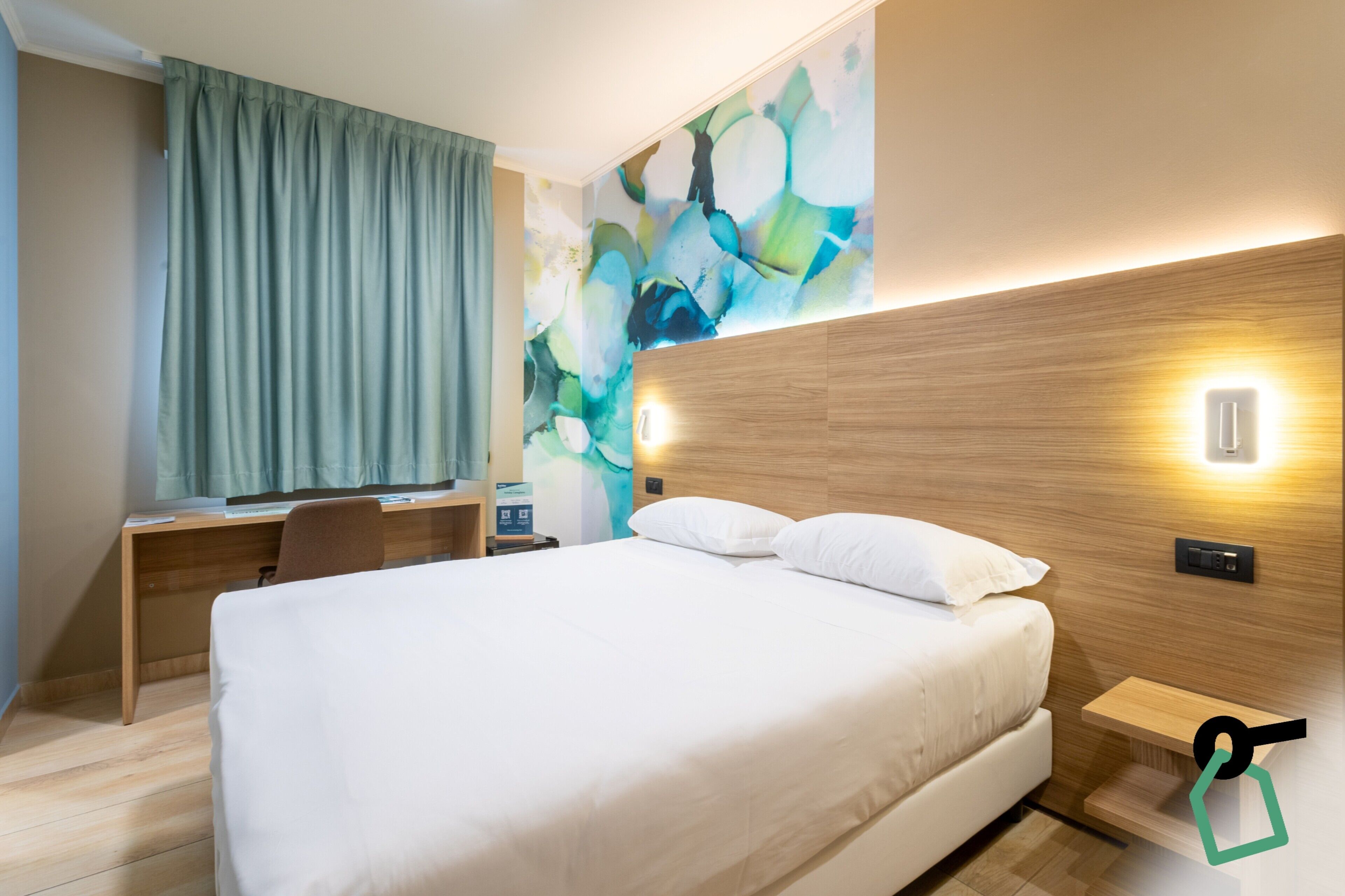 A modern hotel room with a large bed, a desk with a chair, a small table, and a wall-mounted light.