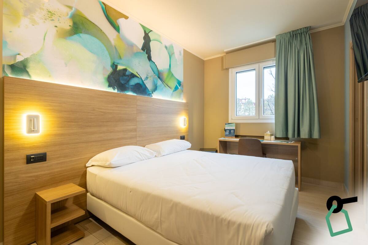 Een hotelkamer met een groot bed, een bureau, een stoel, een klein tafeltje en een raam met gordijnen.