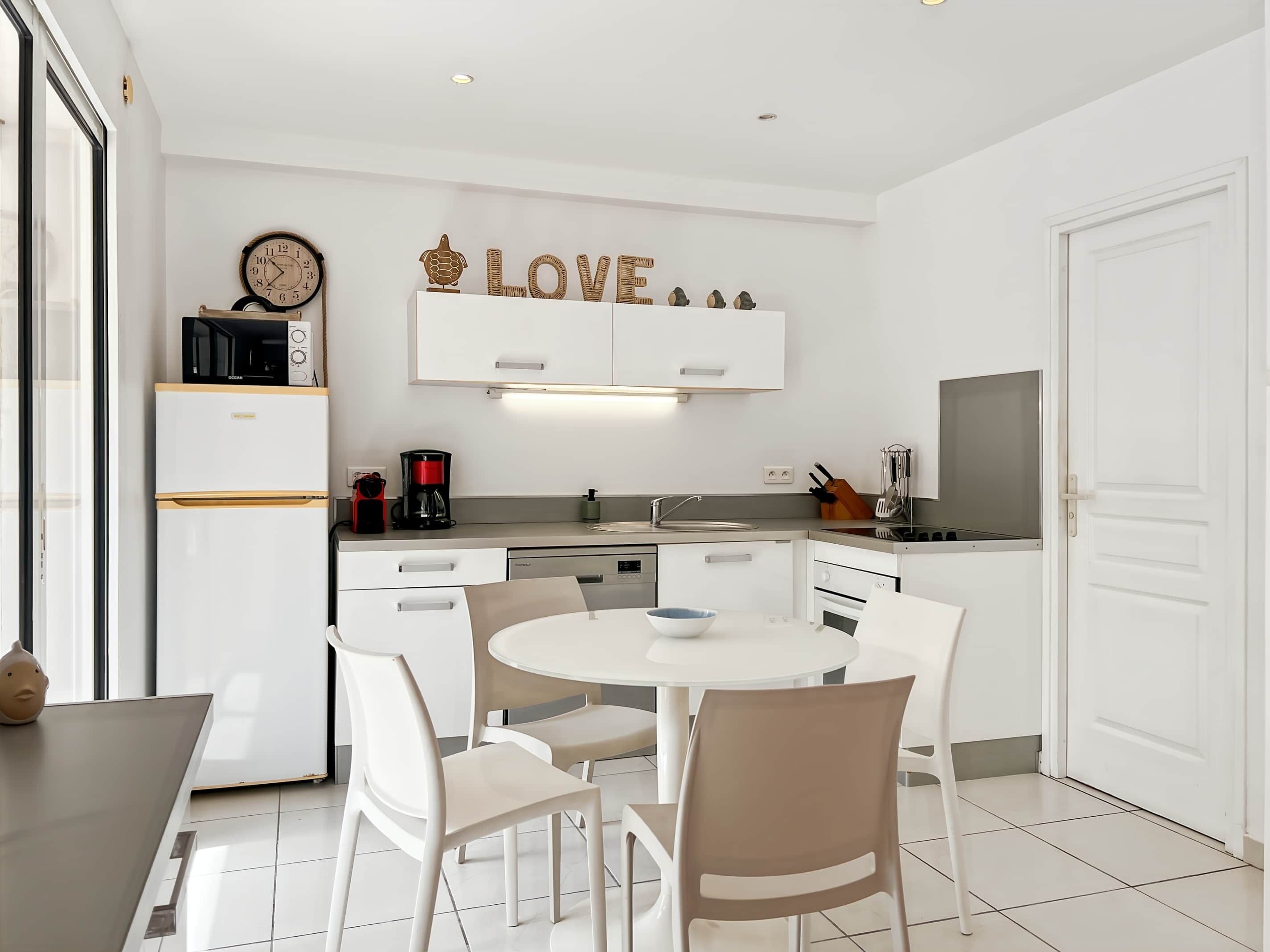 A modern kitchen with white cabinets, a microwave, a coffee maker, and a sink. There is a round table with chairs and a decorative word "LOVE" on the wall.