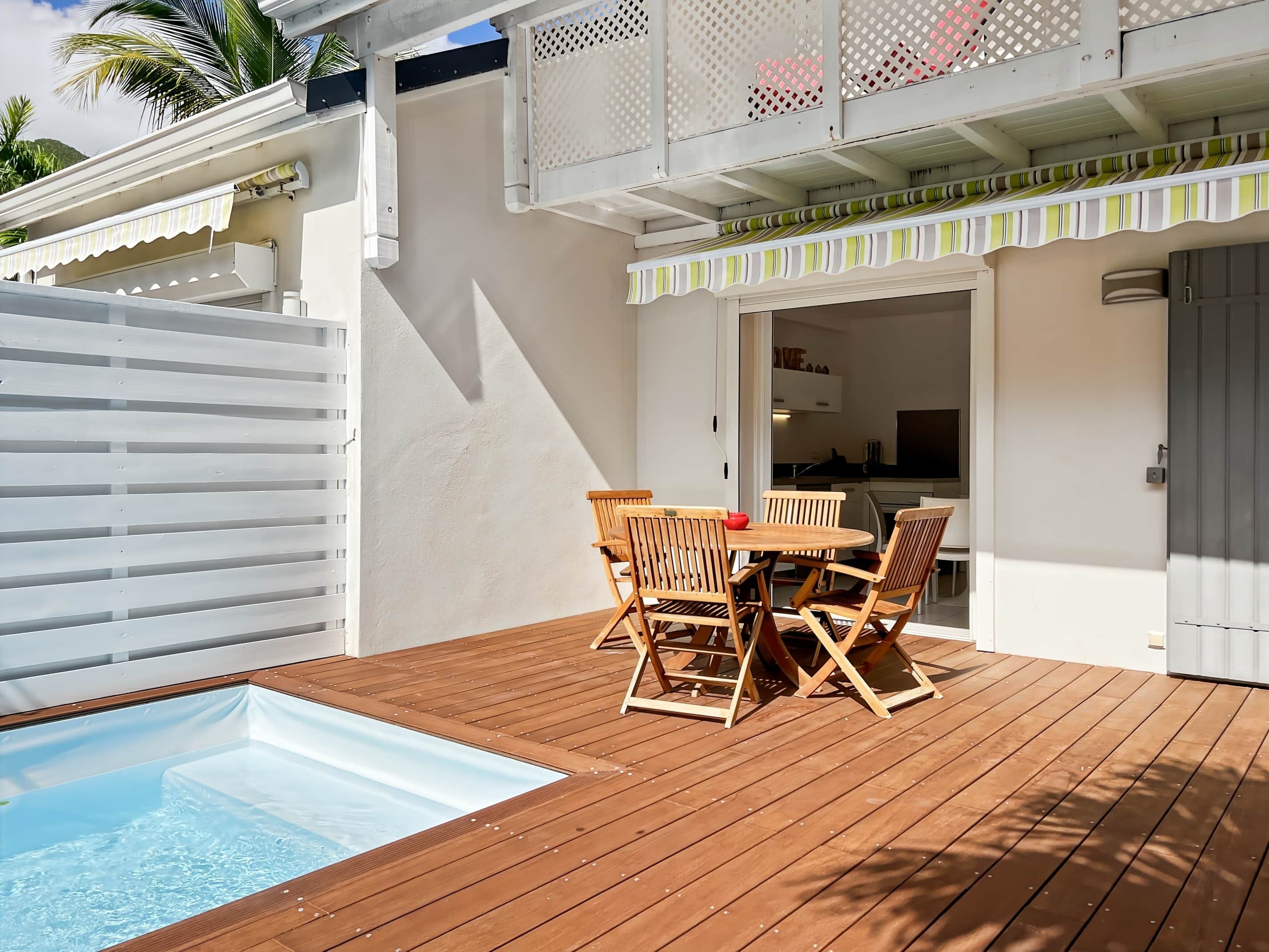 Une terrasse en bois avec une piscine, un coin repas avec des chaises en bois et une cuisine visible à travers une porte ouverte.