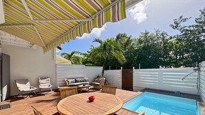 Un jardin avec une piscine, une terrasse en bois, du mobilier d’extérieur et un auvent à rayures.
