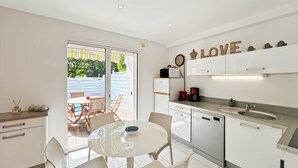 Une cuisine moderne avec des meubles blancs, un coin repas doté d’une table en verre et une vue sur l’extérieur grâce à des portes-fenêtres coulissantes.