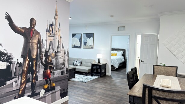 Ein Hotelzimmer mit einer großen Wandmalerei zum Thema Disney, einem Bett mit weißer Tagesdecke, einem grauen Sofa und einem Essbereich mit Tisch und Stühlen.