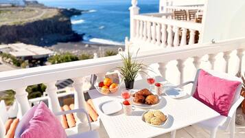 Un balcón con una mesa dispuesta para el desayuno, que incluye cruasanes, un bol de fruta y una copa de vino tinto, con vistas a la costa.