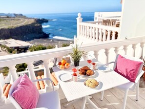 Un balcon avec une table préparée pour le petit-déjeuner, comprenant des croissants, un panier de fruits et un verre de vin rouge, offrant une vue sur la côte.