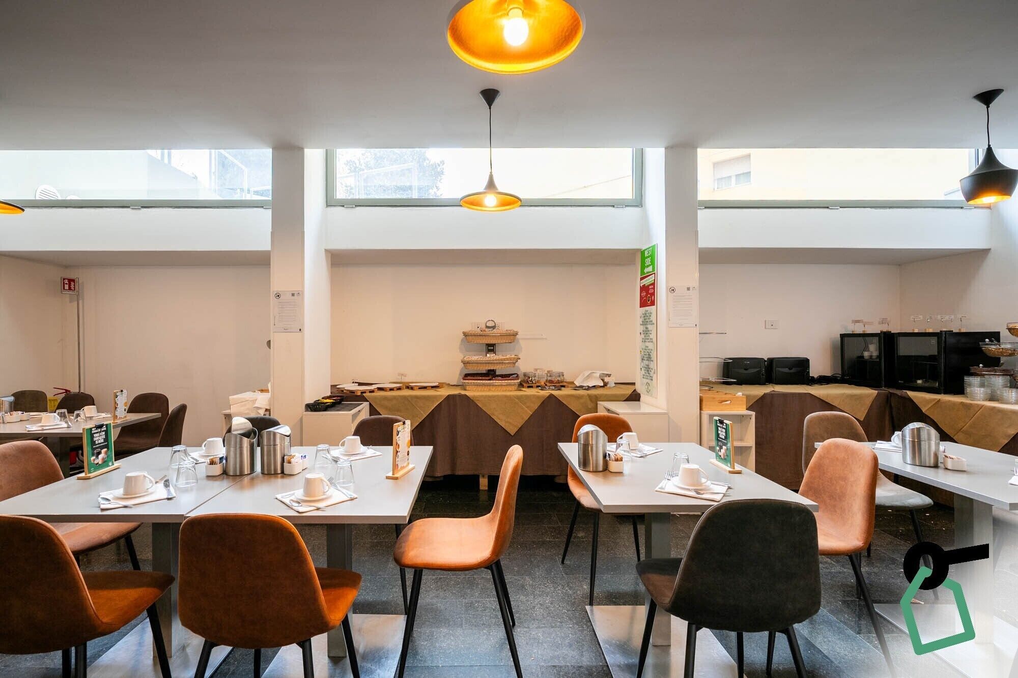 A modern dining area with tables set for meals, overhead lighting, and a view through large windows.