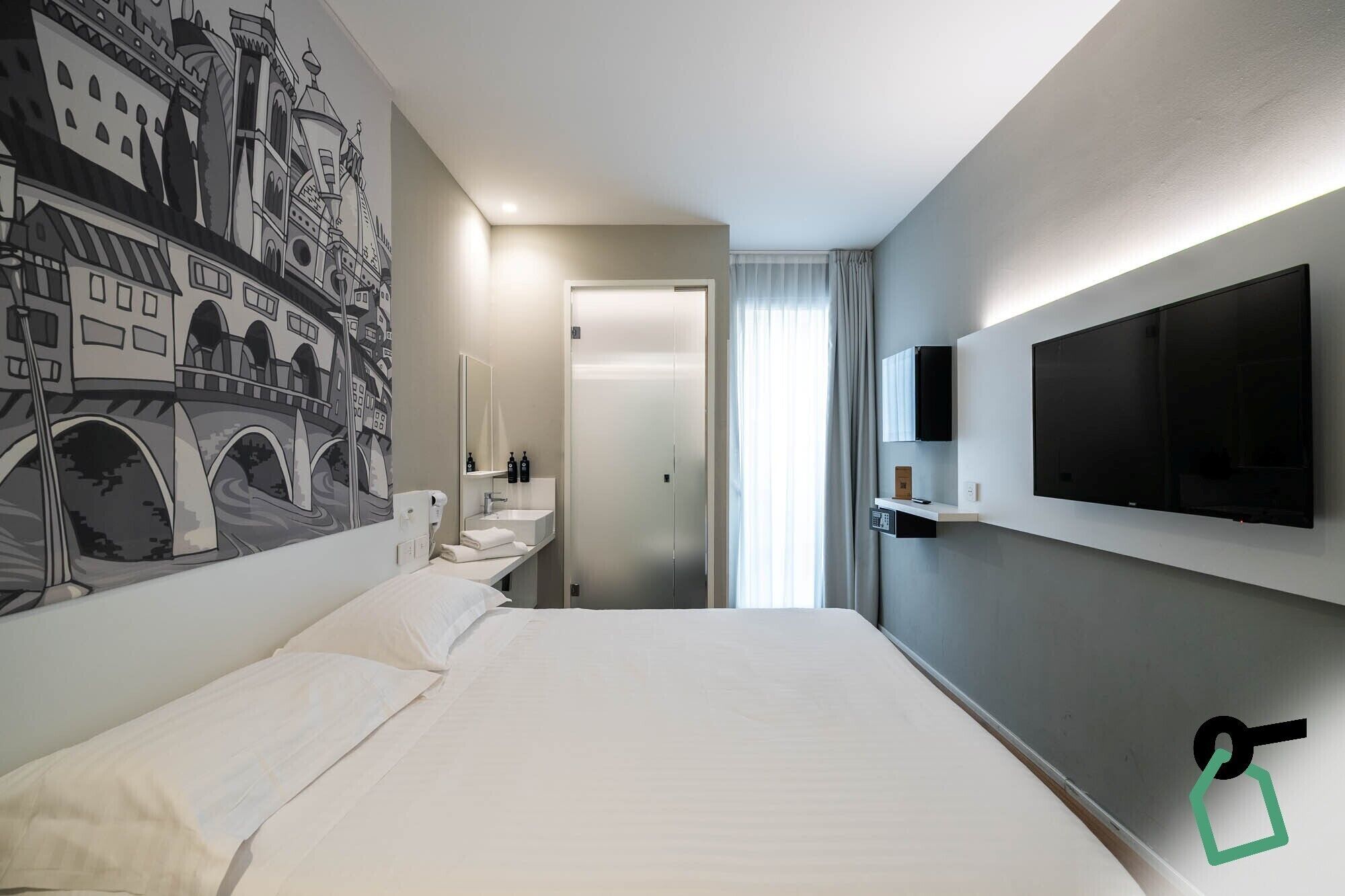 Habitació d'hotel amb un llit gran, un televisor penjat a la paret, un bany amb mirall i rentapel, i un mural d'un paisatge urbà.