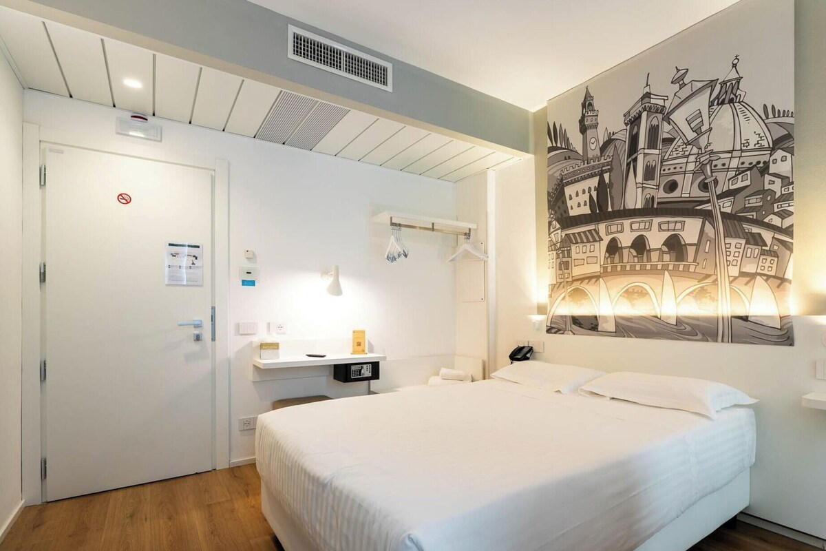 A hotel room with a bed, a desk, a wall-mounted lamp, a painting, and a door with a no-smoking sign.