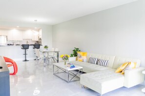 A modern living room with a white sofa, a coffee table, and a dining area with chairs. - Sunny Isles ocean-view 1BR: balcony, pool + gym, steps to beach (Sunny Isles Beach)