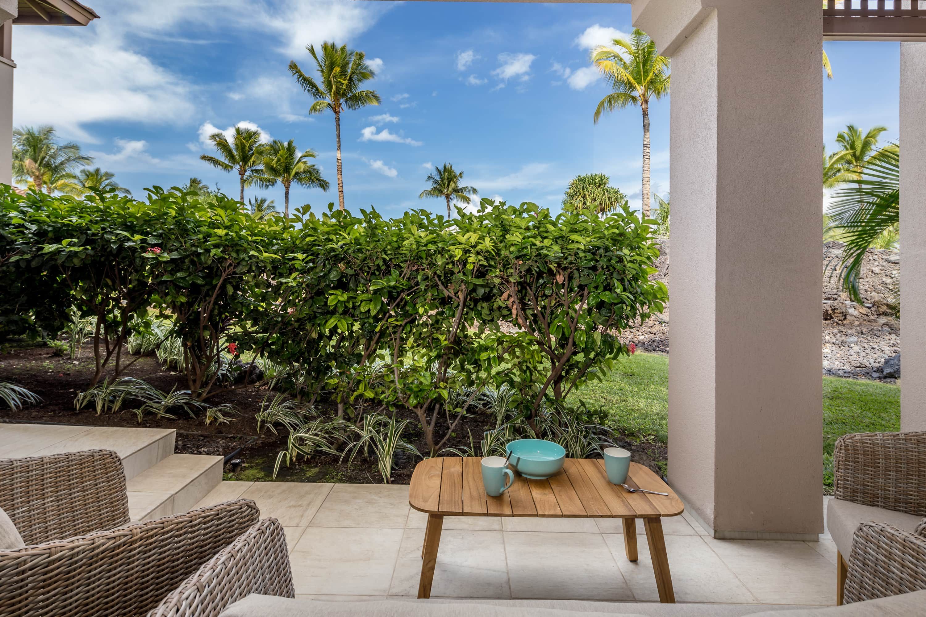 Un patio con mobiliario de mimbre, una mesa de madera y vista a un jardín con palmeras y arbustos.