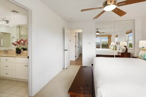 A bedroom with a large bed, a ceiling fan, a nightstand, and a view of the city through the window. - Waikoloa Colony Villas 1702 (Waikoloa Village)