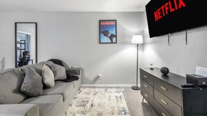 A modern living room with a grey sofa, a flat-screen TV mounted on the wall, a framed poster, a floor lamp, and a dark wooden dresser. - Arlington 1BR | AT&T, Globe Life, 6 Flags, Parking (Arlington)