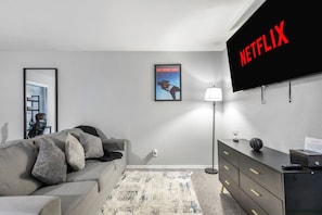 Una sala de estar moderna con un sofá gris, un televisor de pantalla plana montado en la pared, un cartel enmarcado, una lámpara de pie y un aparador de madera oscura.