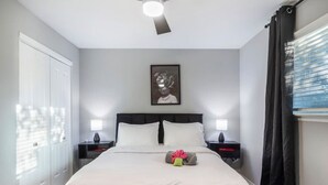 A hotel room with a bed, two bedside tables, a ceiling fan, a framed picture on the wall, and a window with curtains. - Arlington 1BR | AT&T, Globe Life, 6 Flags, Parking (Arlington)