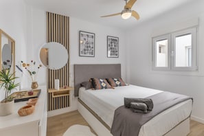 A modern bedroom with a bed, a ceiling fan, a window, and a bedside table with a mirror and decorative items.