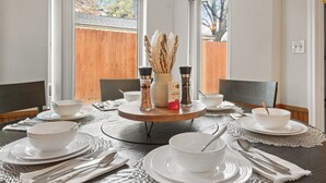 Une table à manger dressée avec des bols, des assiettes et des couverts blancs, entourée de chaises, avec une vue sur une fenêtre et une clôture en bois en arrière-plan.
