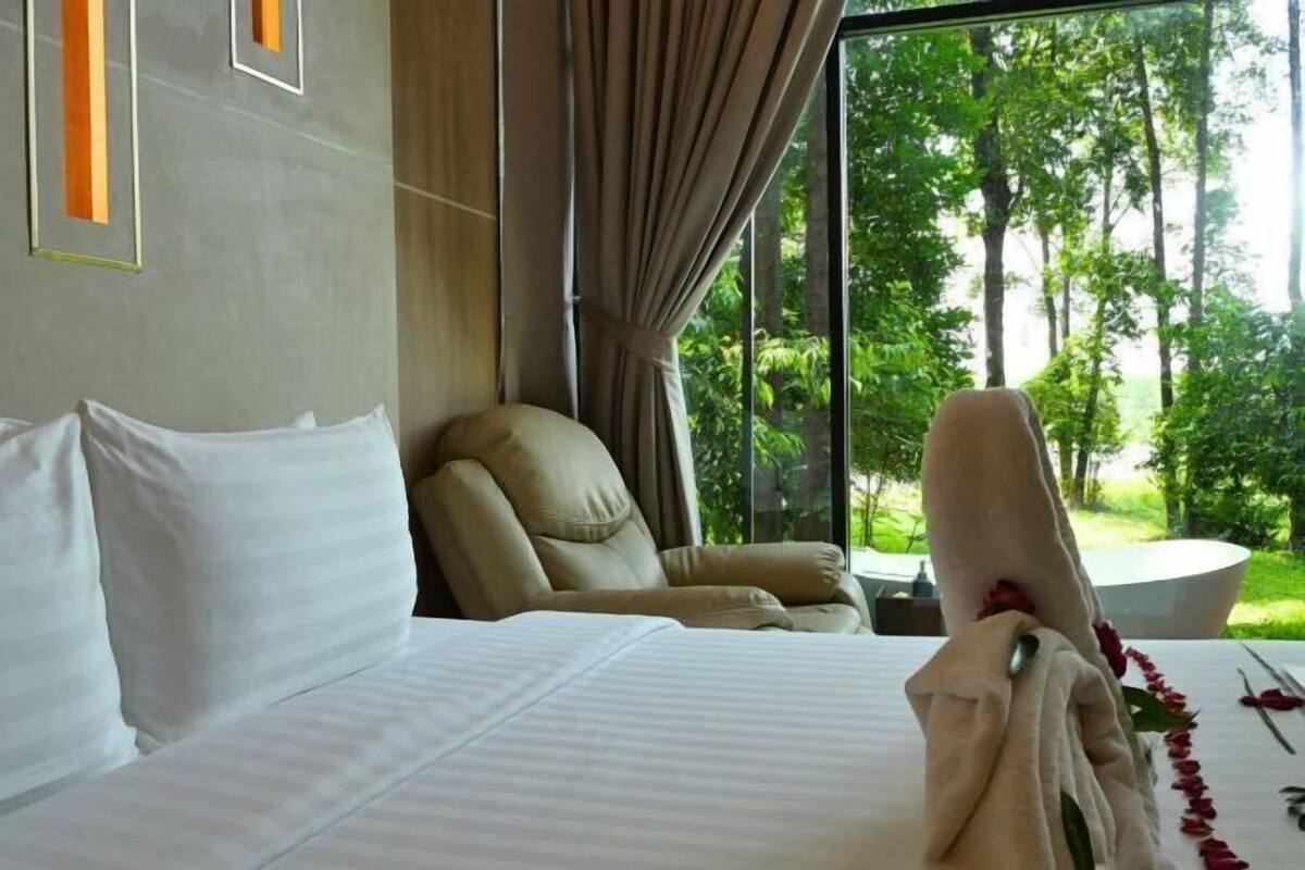 Habitación de hotel con cama, una silla y vistas a árboles a través de grandes ventanales.