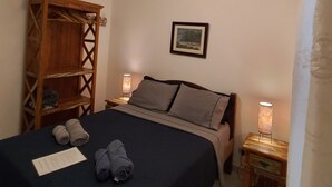 Una cama bien hecha con una colcha oscura, dos almohadas y toallas enrolladas sobre la cama. A la izquierda, una estantería de madera, un cuadro en la pared y dos mesitas de noche con lámparas.