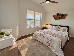 Un dormitorio con cama, una ventana con persianas, ventilador de techo, mesita de noche con lámpara y un adorno de pared.