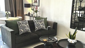 Una sala de estar moderna con un sofá gris, cojines estampados en blanco y negro, una mesita redonda, una mesita auxiliar con una lámpara y una planta en maceta.
