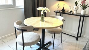 Una mesa redonda de comedor con cuatro sillas, un jarrón con flores, una lámpara y una pequeña mesa con adornos.