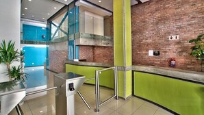 Un vestíbulo con pared de ladrillo, una pared de acento verde, un mostrador de recepción, un ascensor de vidrio y una puerta azul al fondo.