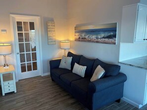 Ein Zimmer mit einem dunkelblauen Sofa, zwei Beistelltischen mit Lampen, einer Glastür und einem gerahmten Schild an der Wand. Außerdem ist ein Strandbild zu sehen.