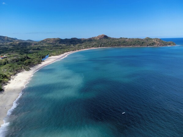 Vista aérea de una zona costera con playa, aguas azules cristalinas y un paisaje montañoso al fondo.