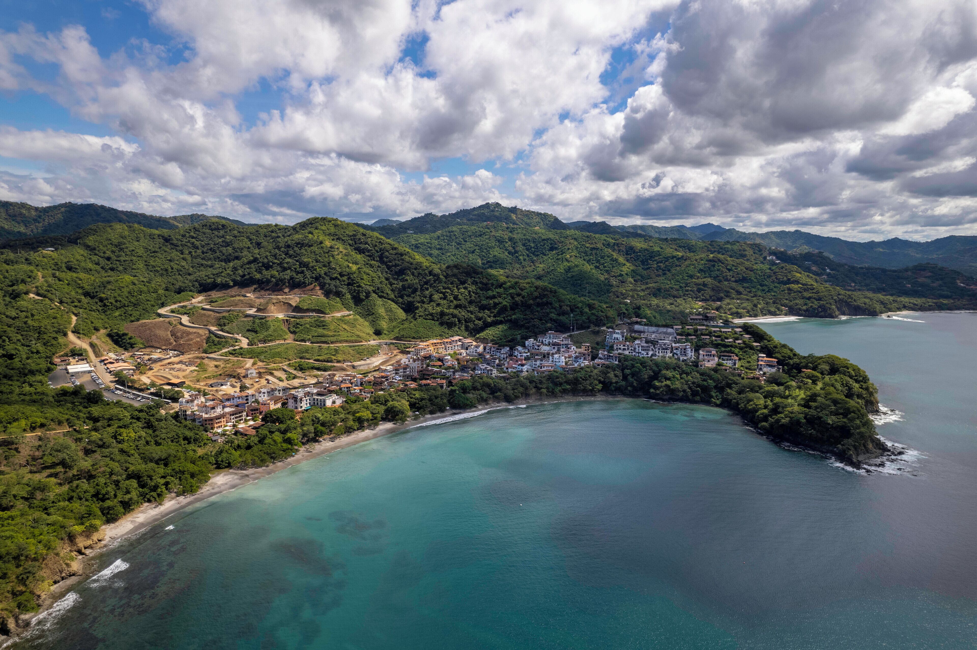An aerial view of a coastal town with a beach, dense greenery, and a mountainous backdrop.