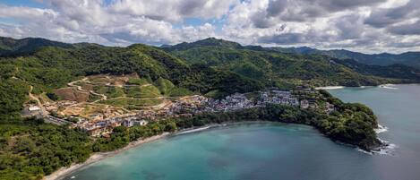 An aerial view of a coastal town with a beach, dense greenery, and a mountainous backdrop.