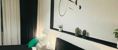 A hotel room with a bed, a nightstand, a wall-mounted shelf with plants, and a decorative wall art piece.